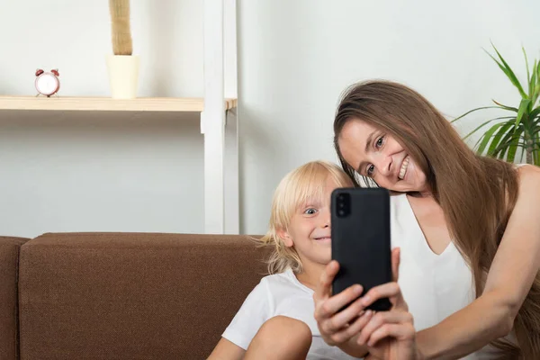 Smiling mom and son take selfie on couch at home. Mom and son take photo on a smartphone