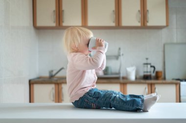 Sarışın çocuk mutfakta oturur ve büyük bardaktan içer. Evde kahvaltı..