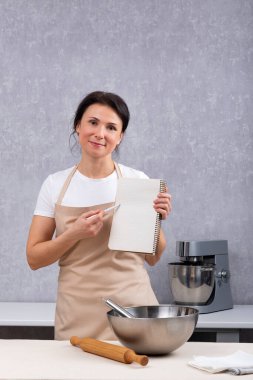 Mutfaktaki önlüklü kadın elinde yemek kitabıyla tarifi açıklıyor. Dikey çerçeve.
