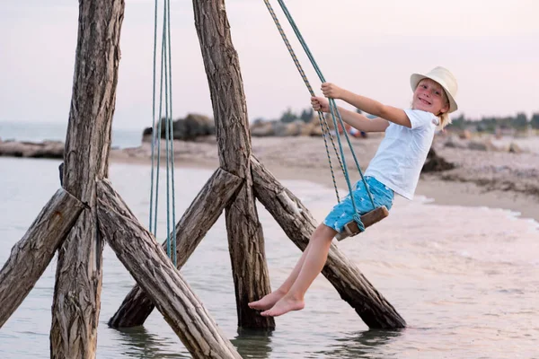 Happy child rides on rope swing over water. Swing by the sea. Holidays with children at sea