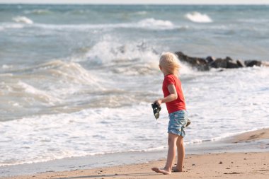 Sarı saçlı çocuk deniz kıyısında yürüyor ve dalgalara bakıyor. Mutlu çocukluk.