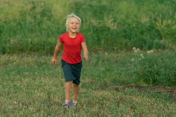 穿着红色T恤的快乐的金发男孩走在绿地上 跑得快 — 图库照片