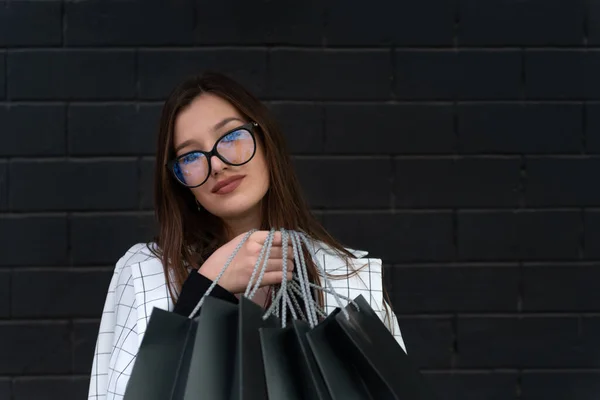 Stylowa Studentka Okularach Trzyma Czarne Torby Zakupy Pod Czarną Ścianą — Zdjęcie stockowe