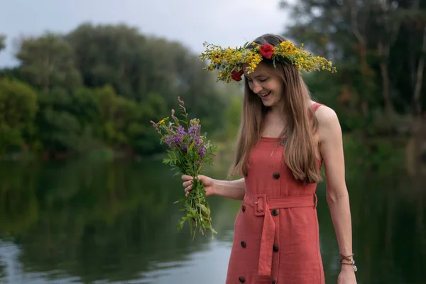 Young woman with wreath on her head with bouquet of wildflowers. River and forest on background. Summer solstice.