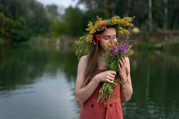 Young girl with wreath on her head holds bouquet of wildflowers on river background. Ivan Kupala holiday.