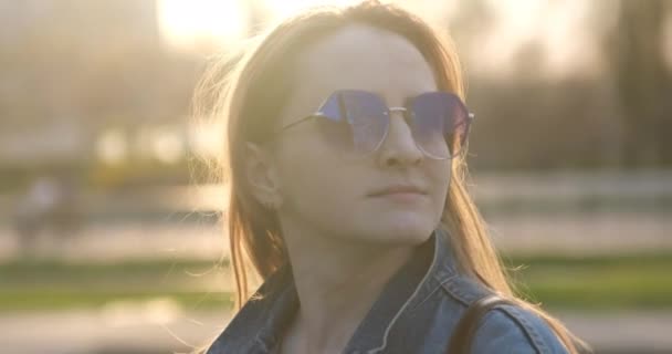 Mujer joven con estilo en gafas de sol endereza su cabello. Puesta de sol urbana sobre fondo — Vídeos de Stock