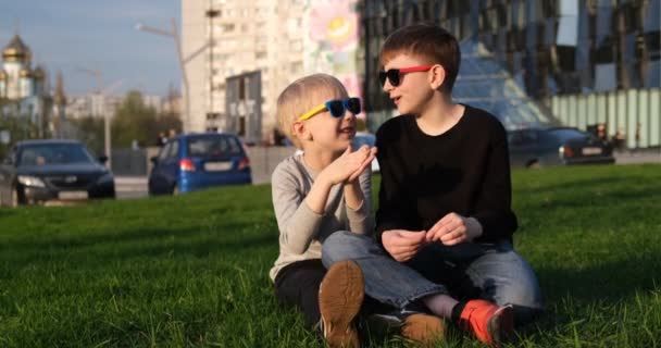 Yeşil çimlerde güneş gözlüklü iki gülen çocuk. Kardeşler bahçede oturarak iletişim kuruyor. Çocukların arkadaşlığı. — Stok video