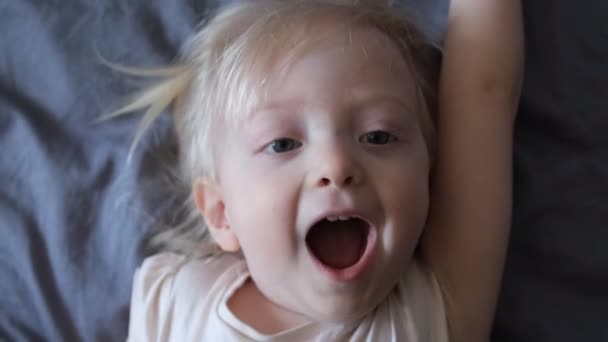 Joyeux bébé blond chante couché sur le lit et glisse ensuite vers le bas. Agréable réveil, enfance heureuse. Vue du dessus. — Video