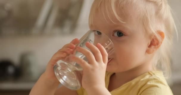Una niña rubia bebe agua de un vaso en la cocina mirando a la cámara y sonriendo — Vídeos de Stock