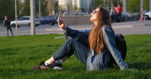 Young woman making selfie on smartphone while sitting on the green lawn. City on the background. Tourist resting on grass — Stock Video