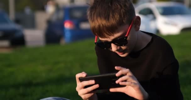 Школьник с помощью смартфона сидит на лужайке. Поколение Z. Мальчик играет по телефону на улице. Современные технологии и дети — стоковое видео