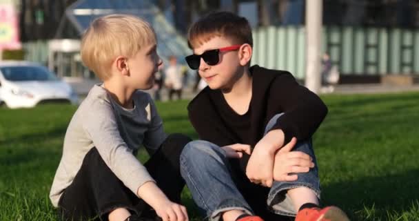 Τα παιδιά συζητούν κάτι ζωντανό. Δύο αγόρια στο γκαζόν με γυαλιά ηλίου διασκεδάζουν κουβεντιάζοντας. Αδέλφια στο γρασίδι. — Αρχείο Βίντεο