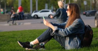 Genç bir kadın yeşil çimlerde otururken akıllı telefon kullanıyor. Otların üzerinde dinlenen mutlu turistler. Şehir ve çocuklar arka planda. 