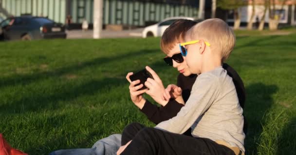 Twee jongens spelen op een smartphone zittend op het gazon. Kinderen met gadget buiten. Vrienden rusten op het gazon. — Stockvideo