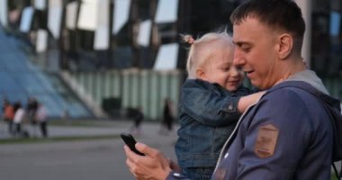 Babası kucağında küçük kızıyla selfie çekiyor. Şirin kız babasına sarılıyor ve seviniyor. Şehirde genç bir aile yürüyor ve video çekiyor.