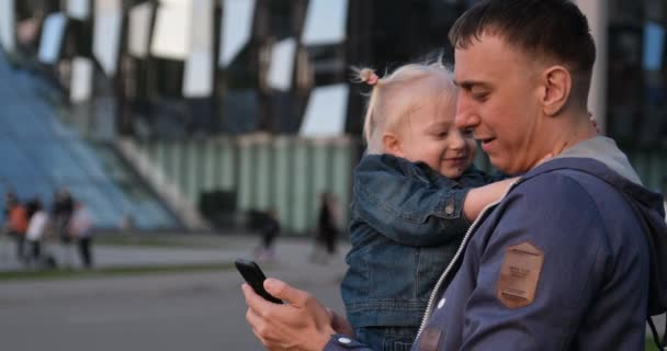 O pai tira uma selfie com a filha nos braços. A filha gira abraça o pai e alegra-se. Jovem família caminha na cidade e filma vídeos — Vídeo de Stock