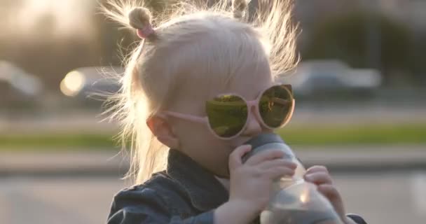 Маленька блондинка в сонцезахисних окулярах п'є воду з пляшки на фоні міста і заходу сонця — стокове відео