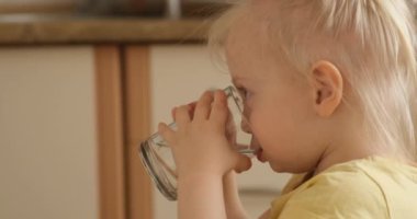 Küçük sarışın bir kız mutfaktaki bardaktan su içiyor kameraya bakıyor ve gülümsüyor.