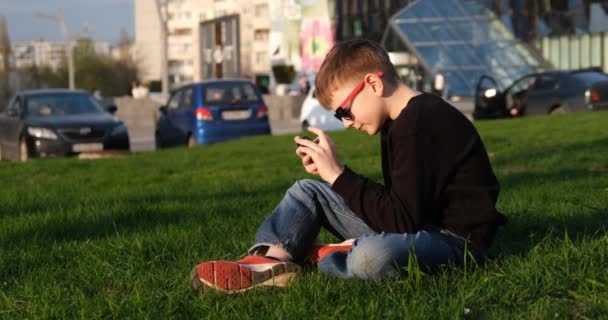 Ο μαθητής παίζει σε ένα smartphone καθισμένος στο γρασίδι. Γενιά Ζ. Το αγόρι χρησιμοποιεί το τηλέφωνο έξω. Σύγχρονη τεχνολογία και παιδιά — Αρχείο Βίντεο