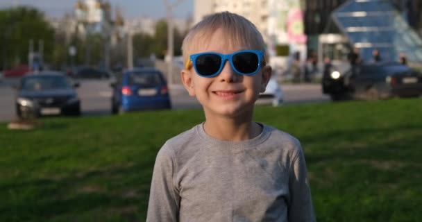 Élégant garçon souriant blond dans des lunettes de soleil se tient sur la pelouse. Le jeune écolier profite des vacances. La ville en arrière-plan — Video