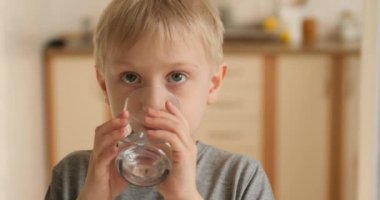 Blond boy drinks water from a glass and shows the class. Joyful child with pleasure drinks water.