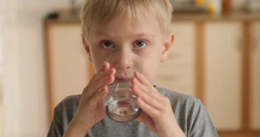 Calm boy drinks water from a glass and shows the class. Baby quenches thirst in the kitchen
