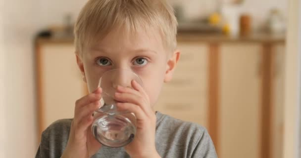 Blonďák pije vodu ze sklenice a ukazuje třídu. Radostné dítě s radostí pije vodu. — Stock video
