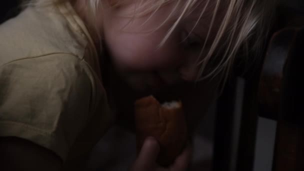 Hungry child hides under a table and eats a piece of bread. Girl eats forbidden food. Harmful gluten food — Vídeo de Stock