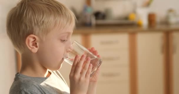 Blond garçon boit de l'eau d'un verre dans la cuisine. L'enfant étouffe la soif. Garçon heureux avec un verre d'eau — Video