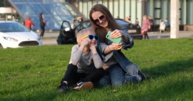 Mom and son laugh and take a selfie on a smartphone sitting on the lawn in the city. Mom and child have fun outdoors. Summer holidays