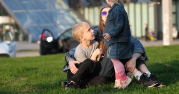 Young mother with two children rests on the lawn. Siblings play with their mom in the fresh air. Family relaxes in the city on the lawn — Vídeo de Stock