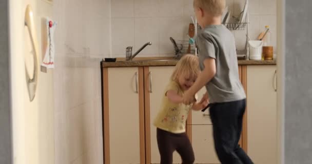 Children dance and jump in the kitchen. Brother and sister have fun together. Blonde sbillings rejoice — Vídeo de Stock
