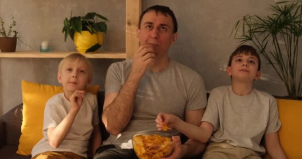 El joven padre con dos hijos pequeños se sienta en el sofá y come papas fritas. Mirando la tele. Luz natural — Vídeo de stock