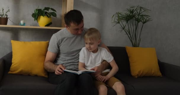 Отец читает книгу сыну, мальчик внимательно слушает. Папа проводит время с ребенком. Дошкольник и папа читают дома на диване — стоковое видео