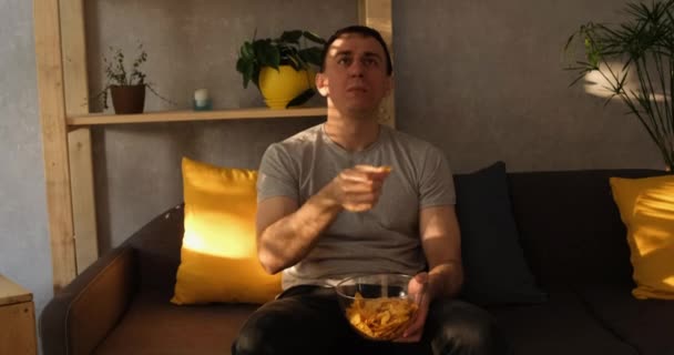 Человек с тарелкой чипсов сидит на диване и ест и смотрит телевизор. Естественное вечернее освещение. Медленное движение — стоковое видео