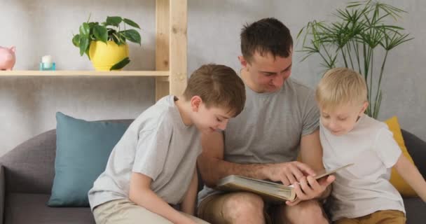 Padre con dos hijos pequeños ve el álbum de fotos de la familia sentado en un sofá en la sala de estar. Valores familiares. Vista frontal de la luz natural — Vídeo de stock