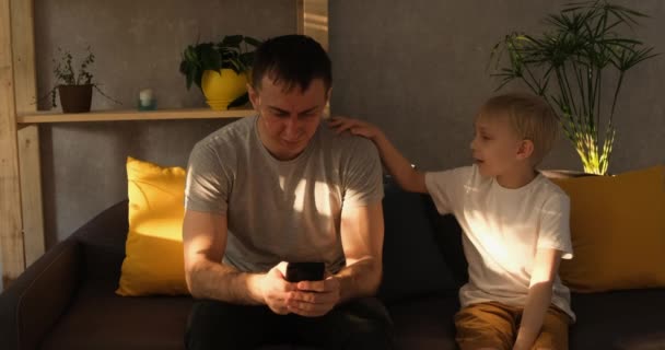 Père est assis sur le canapé et joue dans le smartphone. Le petit fils à côté de lui interfère avec son père et demande de l'attention. Lumière du jour — Video