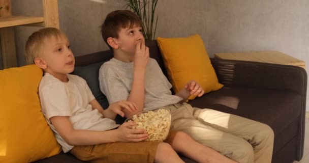 Dva malí kluci jedí popcorn, sedí na gauči a dívají se na televizi. detailní záběr. denní světlo — Stock video