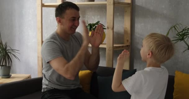 Padre con un pequeño hijo rubio sentarse en el sofá y jugar palmadita un cacke. Aplaudiendo. Vista lateral luz natural del día — Vídeo de stock