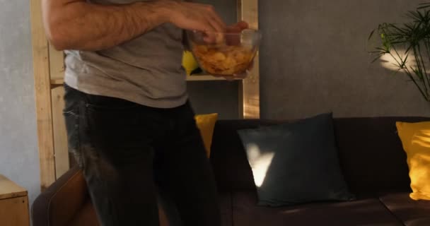 Homme avec une assiette de chips s'adapte et s'assied sur le canapé manger et regarder la télévision. Lumière naturelle du soir. Mouvement lent — Video