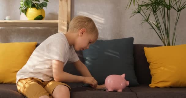 Niño rubio pliega monedas en una alcancía de cerdos sentado en un sofá en la sala de estar. Ahorro para niños — Vídeos de Stock