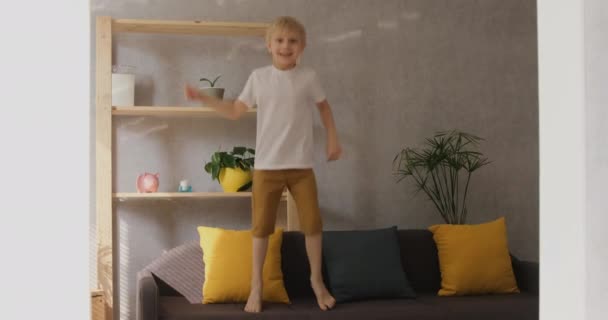 Маленький щасливий блондинка стрибає на дивані у вітальні. Денне світло вид спереду повільний мутація — стокове відео