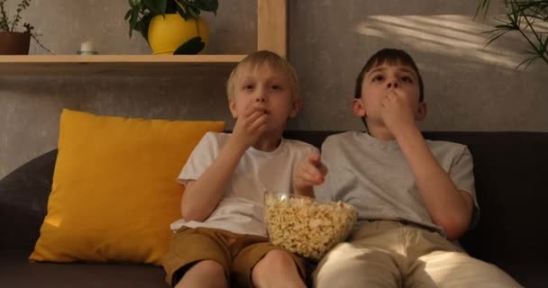 Два маленьких мальчика едят попкорн, сидя на диване и смотря телевизор. крупным планом. daylight — стоковое видео