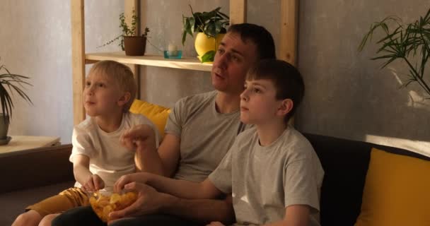 Молодой отец с двумя маленькими сыновьями сидит на диване и ест чипсы. Смотрел телевизор. Естественное освещение — стоковое видео