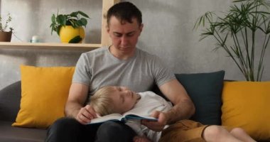 Baba kanepeye oturur ve oğluna babasının kucağında yatan bir kitap okur. Babalık ve çocuklarla vakit geçirme. Gün ışığı