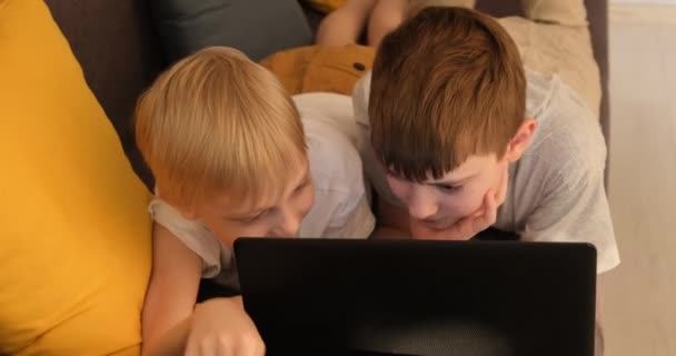 Два мальчика лежат на диване и играют на ноутбуке. Вид спереди. Закрыть — стоковое видео