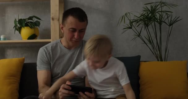 Отец с маленьким сыном играет на смартфоне, сидя на диване. Современное воспитание. Отцы и дети — стоковое видео