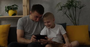 Kanepede oturan akıllı telefondan oynayan küçük bir oğlu olan baba. Modern terbiye. Babalar ve çocuklar