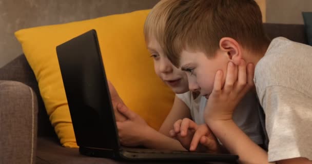 Dois meninos deitados no sofá sorriem e jogam em um laptop. Vista lateral. Fechar — Vídeo de Stock