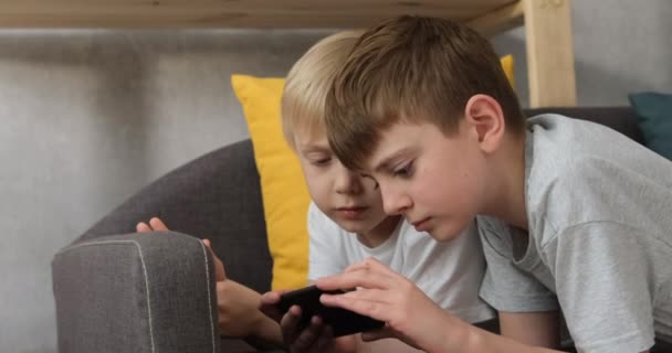 2人の男の子がソファの上に横たわるスマートフォンで遊んでいる。子供やガジェット。昼光サイドビュー — ストック動画